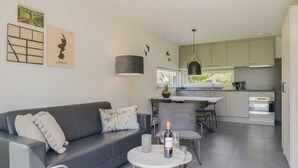 Ett modernt vardagsrum med en grå soffa, ett litet runt bord med en flaska och glas, och en matplats med en grå stol och ett bord.