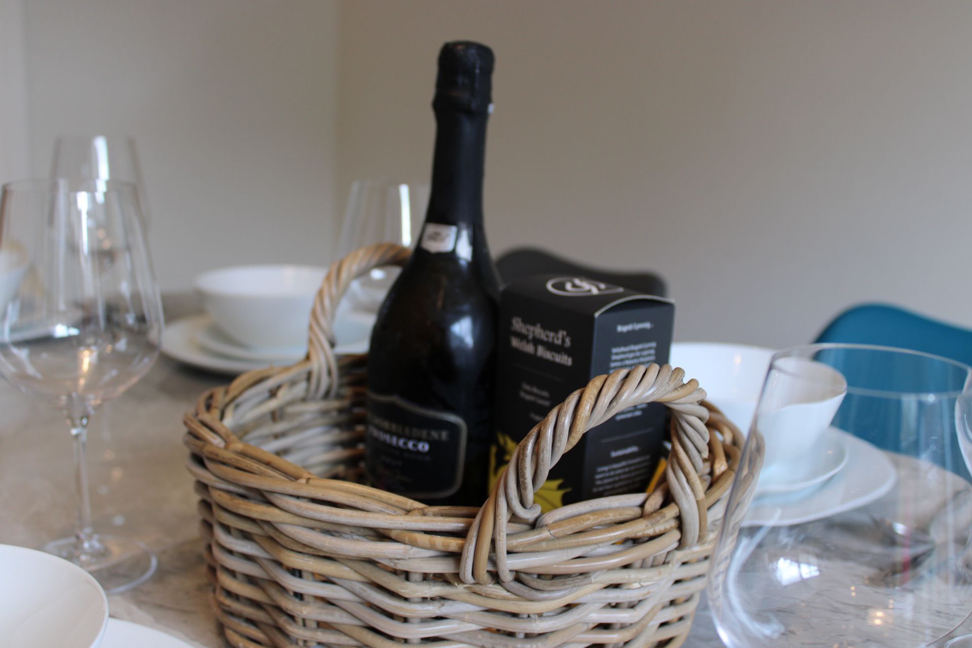 A wicker basket containing a bottle of wine and a box of biscuits on a table with glasses and plates.