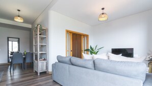 A modern living room with a grey sofa, a wooden bookshelf, a TV, and a dining area with chairs and a table.
