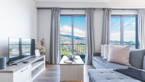 A modern living room with a large TV, a sofa, a coffee table, and a view of a cityscape through large windows.
