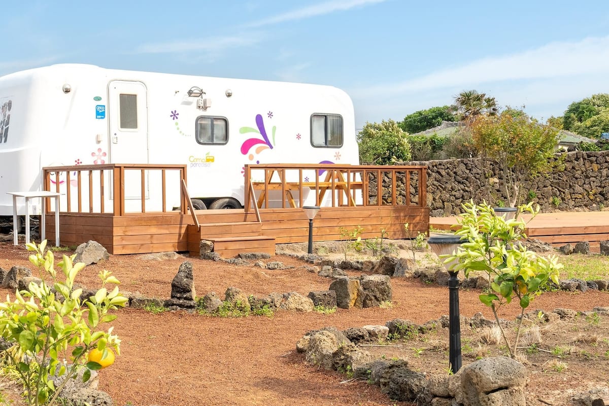A white RV with a wooden deck and steps leading to a door, surrounded by a rocky landscape with sparse vegetation.
