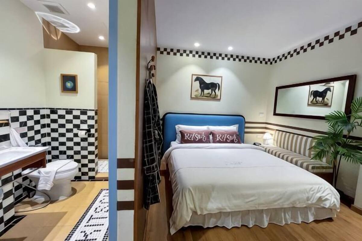 A hotel room with a checkered wall, a bed with a blue headboard, a bedside table, a mirror, and a plant.