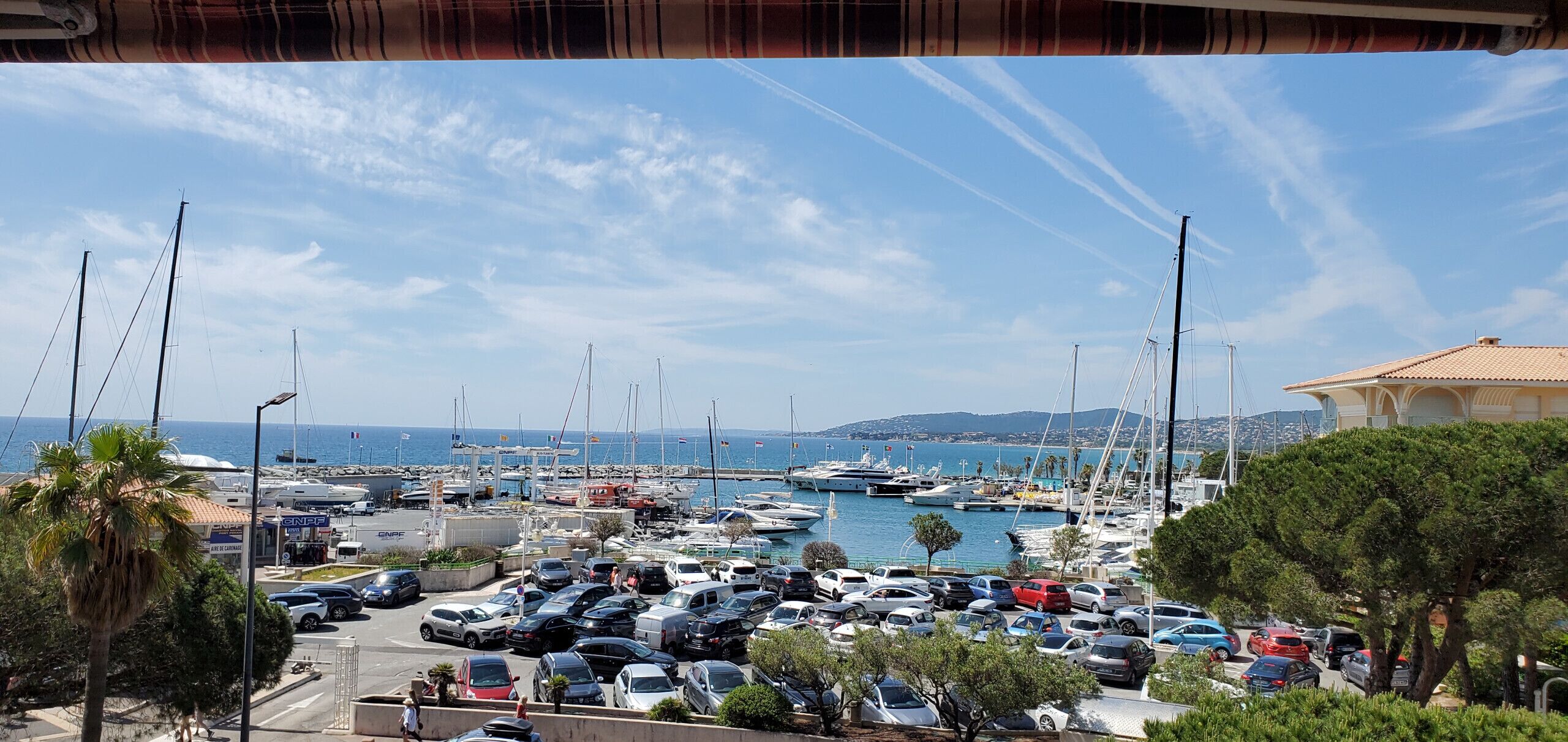A marina with numerous boats docked, a parking lot with cars, and a clear view of the sea and distant hills.