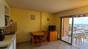 A compact living space with a kitchen area, a dining table with chairs, a wooden dresser, a TV, and a balcony with a view of a marina. - LE CAPITOLE, Sea view, Studio cabin 27m2 waterfront, 2 adults, 2 children, air conditioning and garage (Fréjus)
