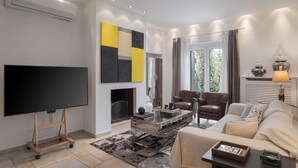 A modern living room with a flat-screen TV, a fireplace, comfortable seating, and a large window with curtains. - Lussuosa Villa Glyfada (Glyfada)