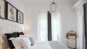 A neatly made bed with white and black pillows, a grey blanket, and a white headboard. There are three framed pictures on the wall, a chandelier, and a wooden bedside table with a lamp. - Lussuosa Villa Glyfada (Glyfada)