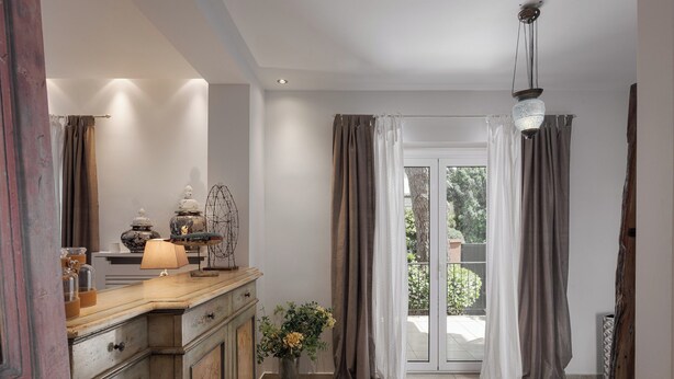 A room with a wooden cabinet, a lamp, and a vase with flowers. There is a sliding glass door leading to an outdoor area. - Lussuosa Villa Glyfada (Glyfada)