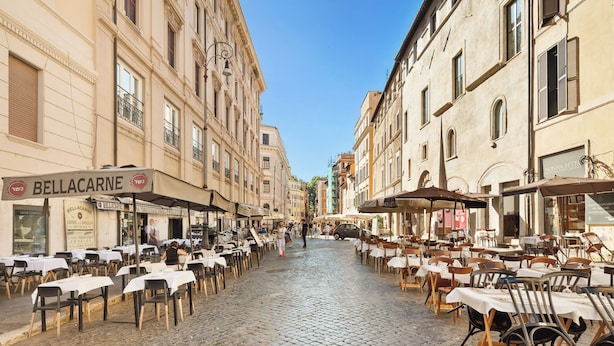 Eine Kopfsteinpflasterstraße gesäumt von Cafés mit Außenbestuhlung, ein klarer, blauer Himmel und historische Gebäude.
