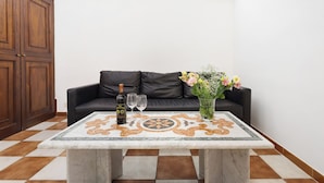 Ein Wohnzimmer mit Schachbrettmuster-Boden, einer schwarzen Ledersofa, einem Marmor-Couchtisch, einer Weinflasche, zwei Weingläsern und einer Vase mit Blumen.