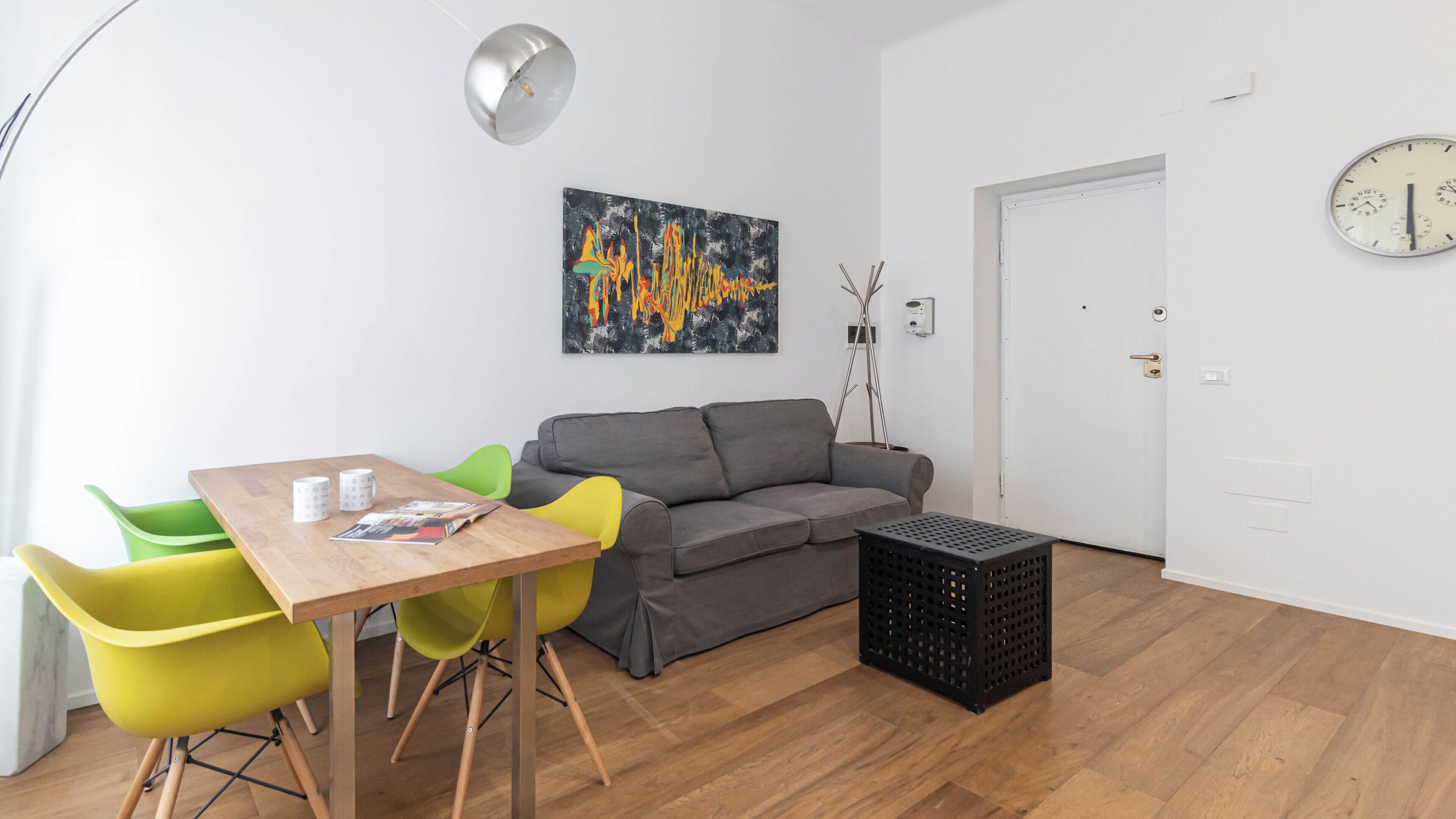 Una sala de estar moderna con una mesa de madera, sillas verdes y amarillas, un sofá gris, un cofre negro y un reloj de pared.