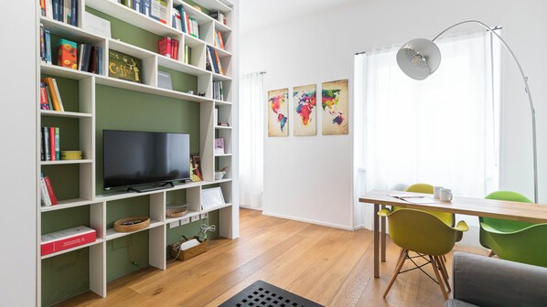 Une pièce comprenant une bibliothèque, un téléviseur à écran plat, une table à manger avec des chaises et une fenêtre avec des rideaux.
