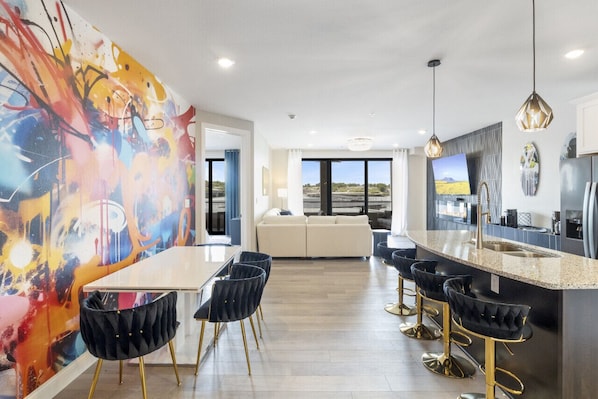 Contemporary kitchen with a central island, sink, and bar stools, featuring stainless steel appliances and a large window with an outdoor view.