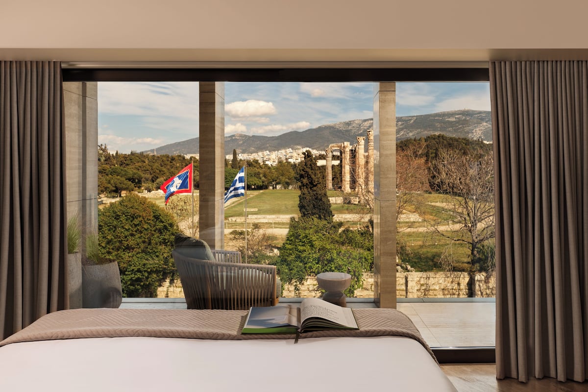 A bedroom with a large window overlooking ancient ruins and flags, a bed, and a reading area with a book.