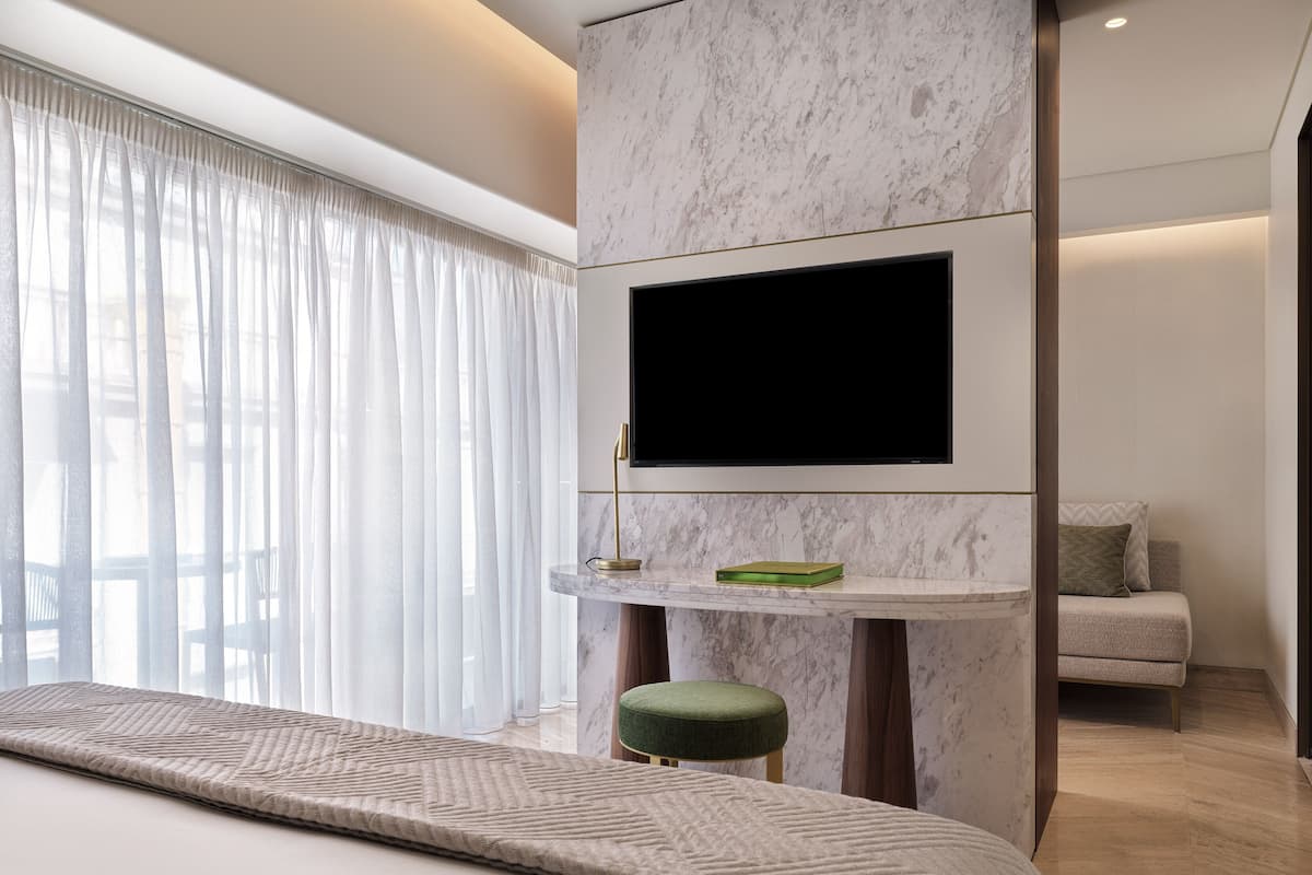 A modern hotel room with a large bed, a flat-screen TV mounted on a marble wall, a small green ottoman, and a seating area with a cushioned bench.