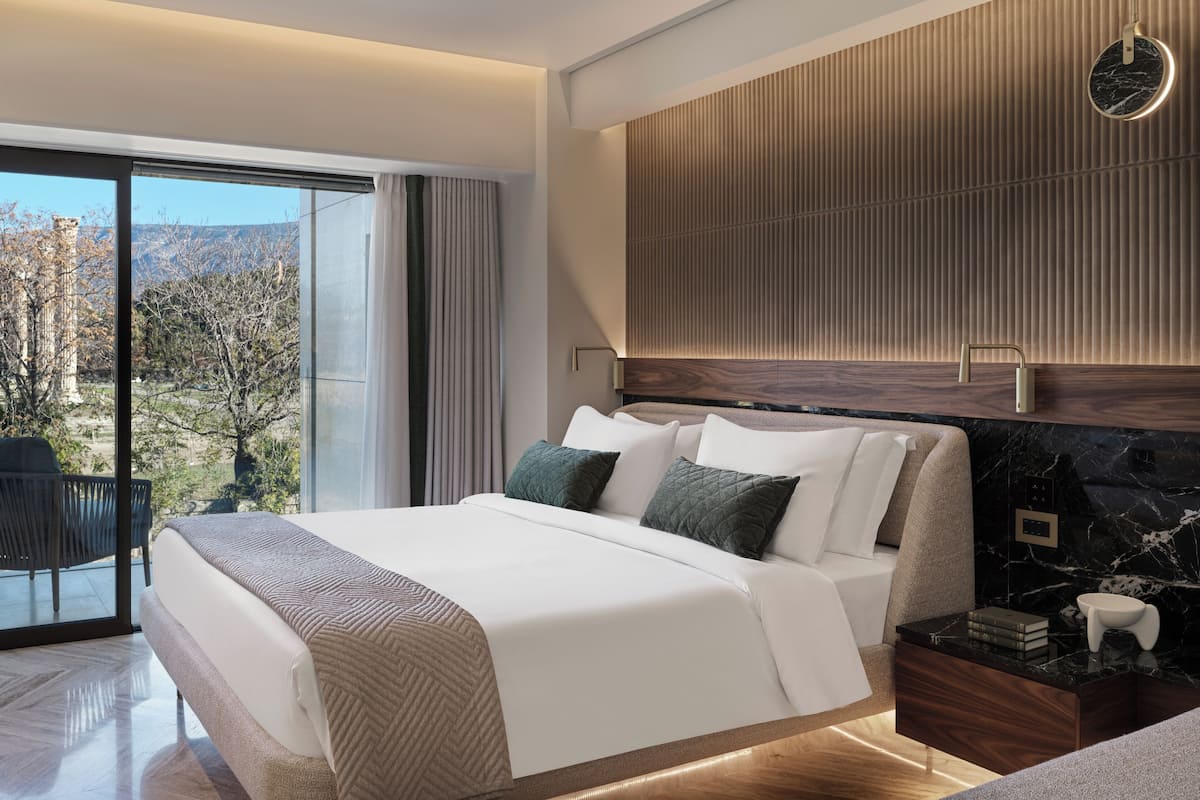 A modern hotel room with a large bed, a wooden headboard, a marble nightstand, and a view of the outdoors.
