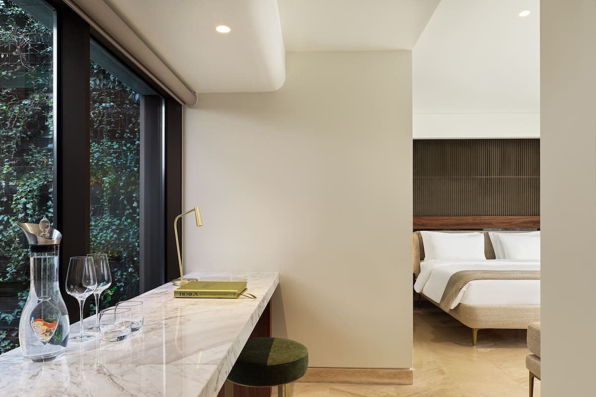 A modern hotel room with a marble countertop, a green upholstered stool, a bed with white linens, and a view of greenery through large windows.