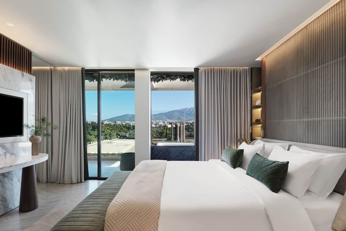 A modern hotel room with a large bed, a TV mounted on the wall, and a view of the city and mountains through large windows.