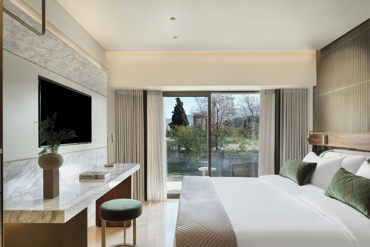 A modern hotel room with a large bed, a desk with a chair, a TV, and a view of the outdoors.