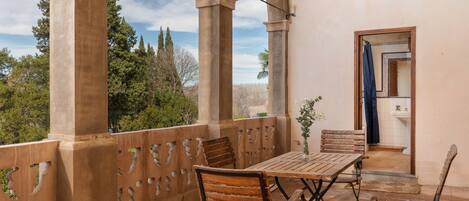 Un balcon avec une table et des chaises, une vue sur les arbres et une porte donnant sur une salle de bain.