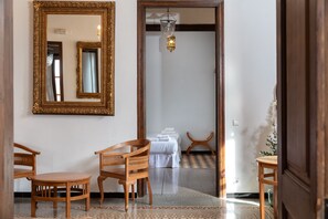 A room with a large ornate mirror, a wooden chair, a small round table, and a bed with white linens.