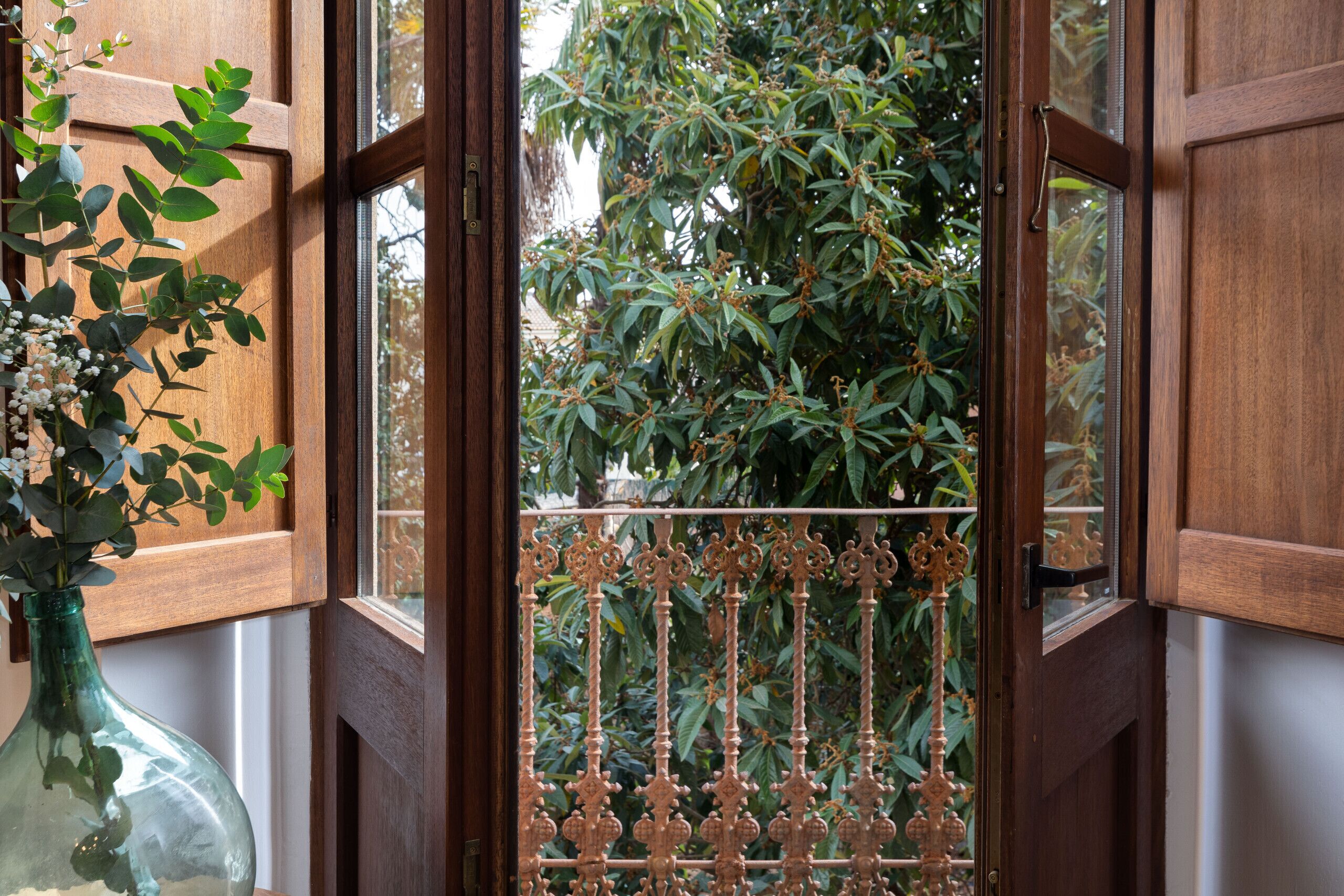 Une porte en bois ouverte, surmontée d’un portail en fer forgé, offre une vue dégagée sur un arbre verdoyant à l’extérieur. Sur la gauche, on aperçoit un vase en verre transparent contenant des fleurs blanches.