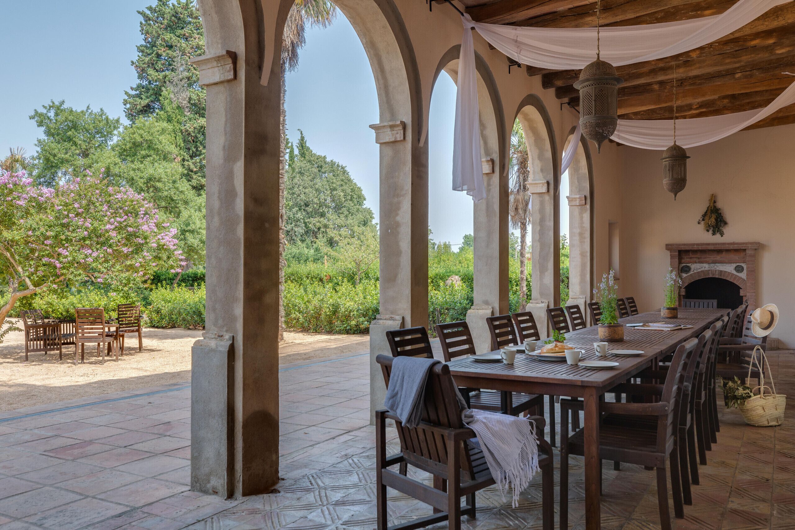 An outdoor patio with a long wooden table and chairs, surrounded by stone columns, with a fireplace and greenery in the background.