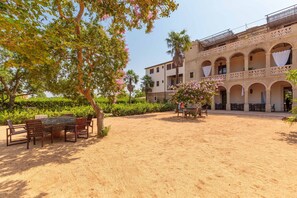 Une cour avec une table et des chaises, entourée de verdure et bordée d’un bâtiment doté de nombreuses arcades et de balcons.