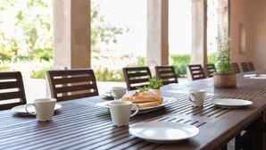 Een houten tafel met borden, bekers en een brood, in een overdekt buitengebied met stoelen en een potplant.