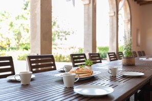 A wooden table with plates, cups, and a loaf of bread, set in a covered outdoor area with chairs and a potted plant. - Flateli. Casa Indiana (Bordils)