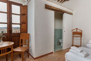 A room with a wooden chair, a small table with a vase, a bed with white linens, and a view through an open door to a bathroom.
