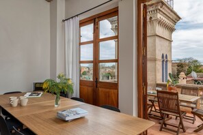 A dining area with a wooden table, chairs, a vase with flowers, and a view of buildings through a large window.