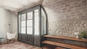 A room with a stone wall, a wooden bench, a white chair, and a large arched window.