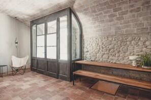A room with a stone wall, a wooden bench, a white chair, and a large arched window.