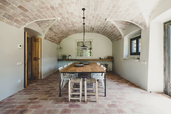 A spacious dining area with a long wooden table, white chairs, and a brick-patterned floor. - Flateli. Casa Oliver (Cervià de ter)