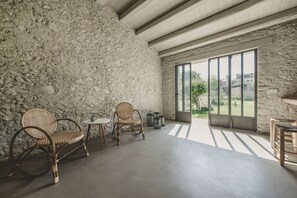 Une pièce avec un mur en pierre, des chaises en osier, une petite table et une vue dégagée sur l’extérieur grâce à de grandes fenêtres.