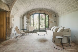 Quarto com teto de pedra, porta de madeira, sofá branco, mesa redonda, dois bancos altos e uma cadeira.