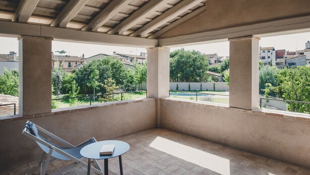 A covered outdoor area with a table and chair, large windows, and a view of greenery and buildings.