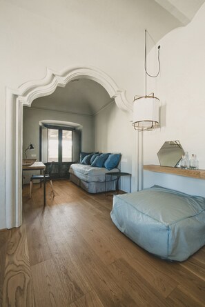 A room with a wooden floor, a blue bean bag, a desk with a lamp, and a window with curtains.