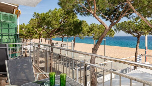 Een balkon met een tafel en stoelen, met uitzicht op een strand met bomen en de zee.