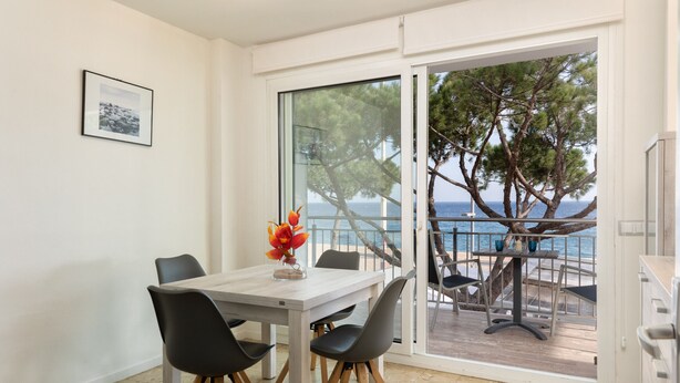 A dining area with a table and chairs, a vase with flowers, and a view of the sea through a glass door. - Flateli. PDA Vista Mar 2 (Platja d´Aro)