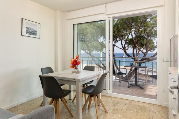 Un comedor con mesa y sillas, un florero con flores y vista al mar a través de un ventanal.