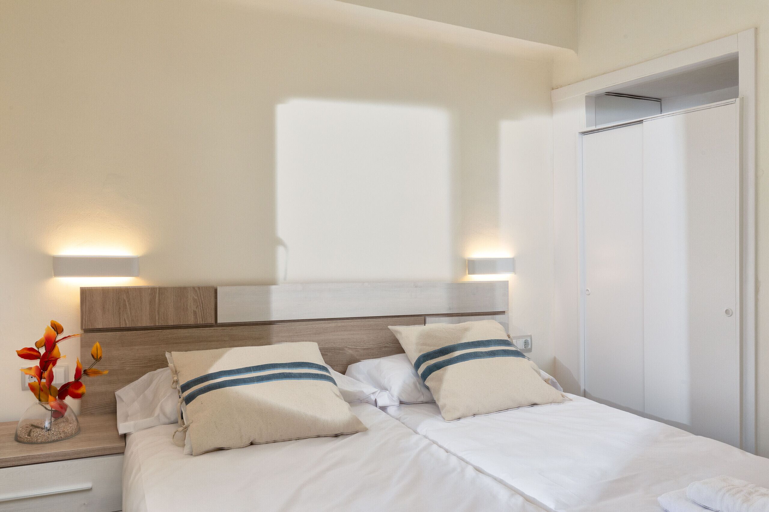 Habitación de hotel con cama, lámparas de noche, mesita de noche, un jarrón con flores y un armario.