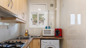 A kitchen with a gas stove, microwave, washing machine, and a window with a view of trees. - Flateli. PDA Vista Mar 2 (Platja d´Aro)