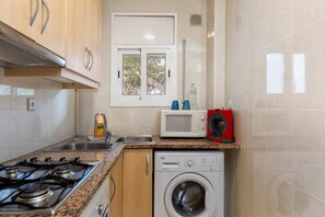 A kitchen with a gas stove, microwave, washing machine, and a window with a view of trees.