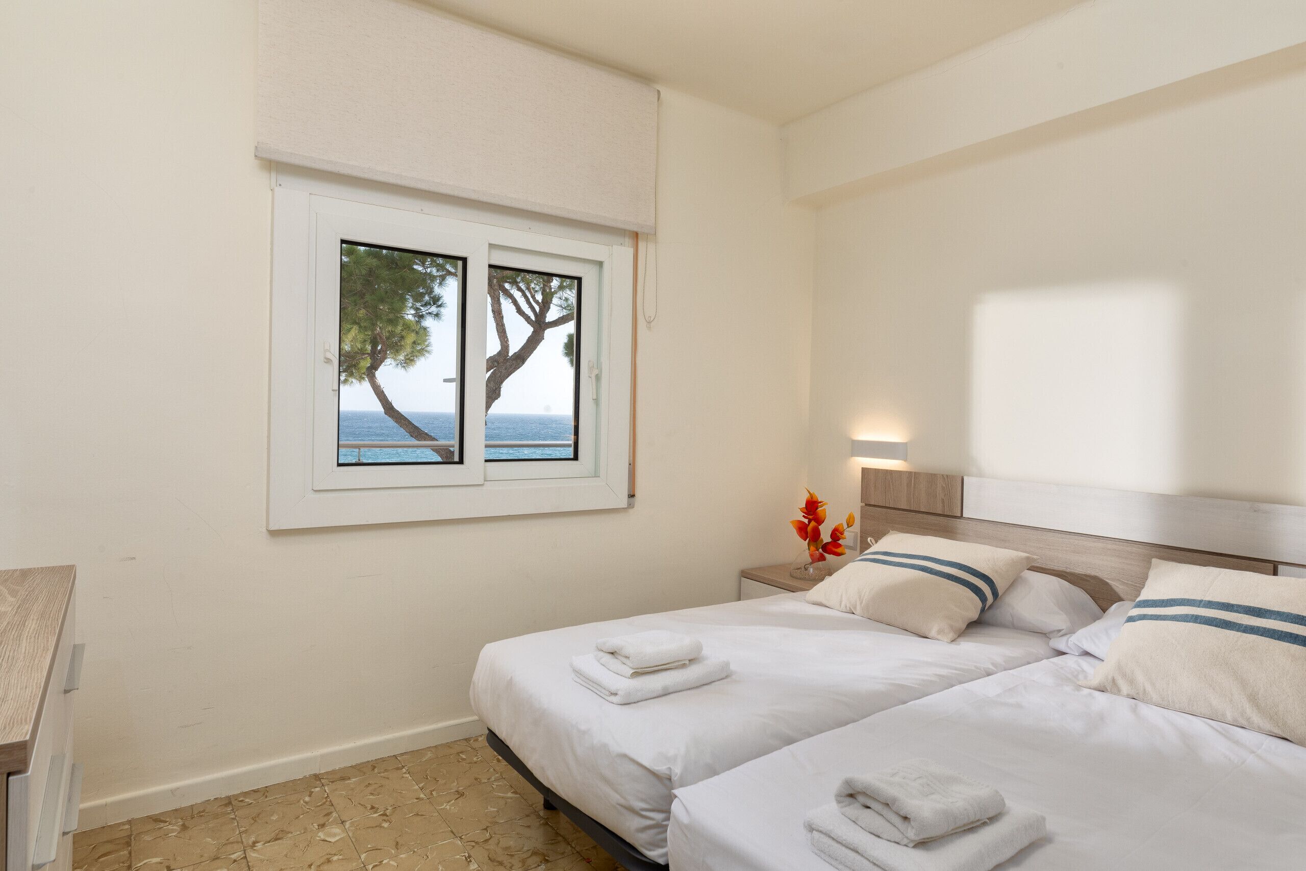 A hotel room with two beds, a window with a view of the sea, and a nightstand with a vase of flowers.