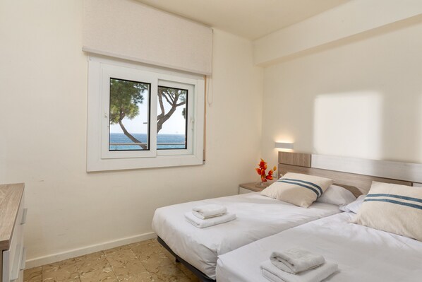 A bedroom with two beds, a window with a view of the sea, and a nightstand with a vase of flowers.