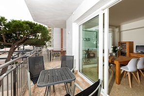 Un balcón con una mesa y sillas, una puerta de vidrio que da a una habitación con televisión y armarios, y un árbol al fondo.