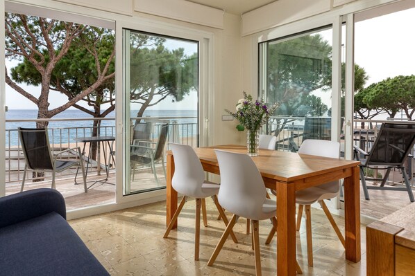 A dining area with a wooden table and white chairs, large glass doors leading to a balcony with a view of the sea and trees.
