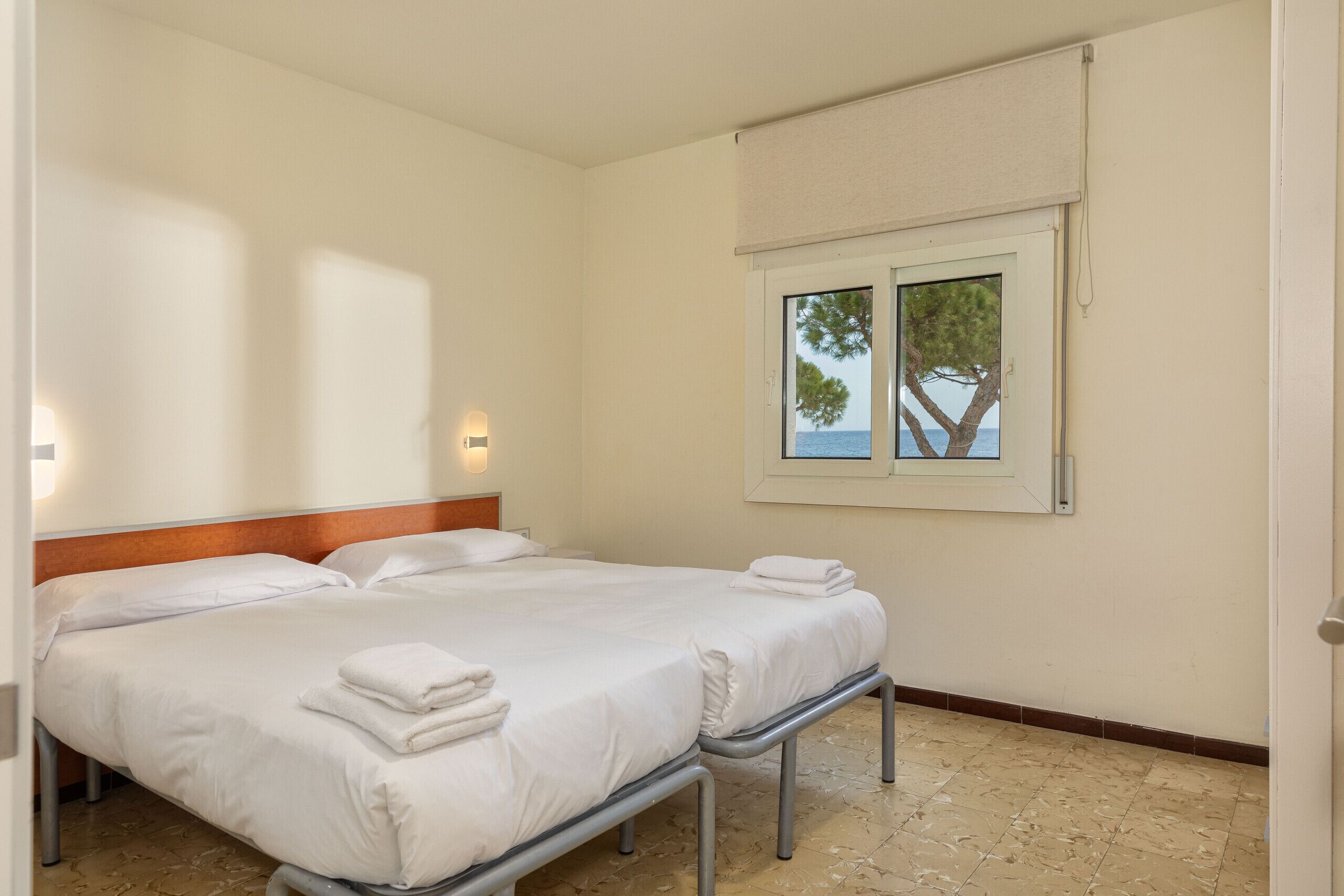 Un lit double avec du linge de lit et des serviettes blanches, une tête de lit en bois, une fenêtre donnant sur les arbres et la mer, et un luminaire fixé au mur.