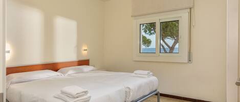 A double bed with white linens and towels, a wooden headboard, a window with a view of trees and the sea, and a wall-mounted light fixture.